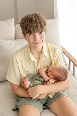 Older brother sitting on a bed, gently holding his newborn baby brother in a sage green outfit.