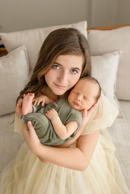 Big sister sitting on a bed, cradling her newborn baby brother in a sage green outfit