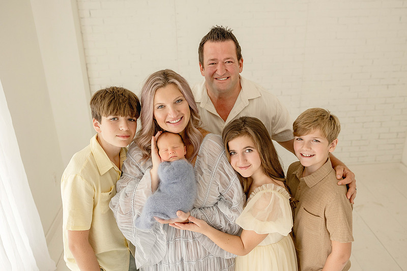 Mother holding her newborn baby boy, with father standing behind her and three older siblings gathered around them for a family portrait
